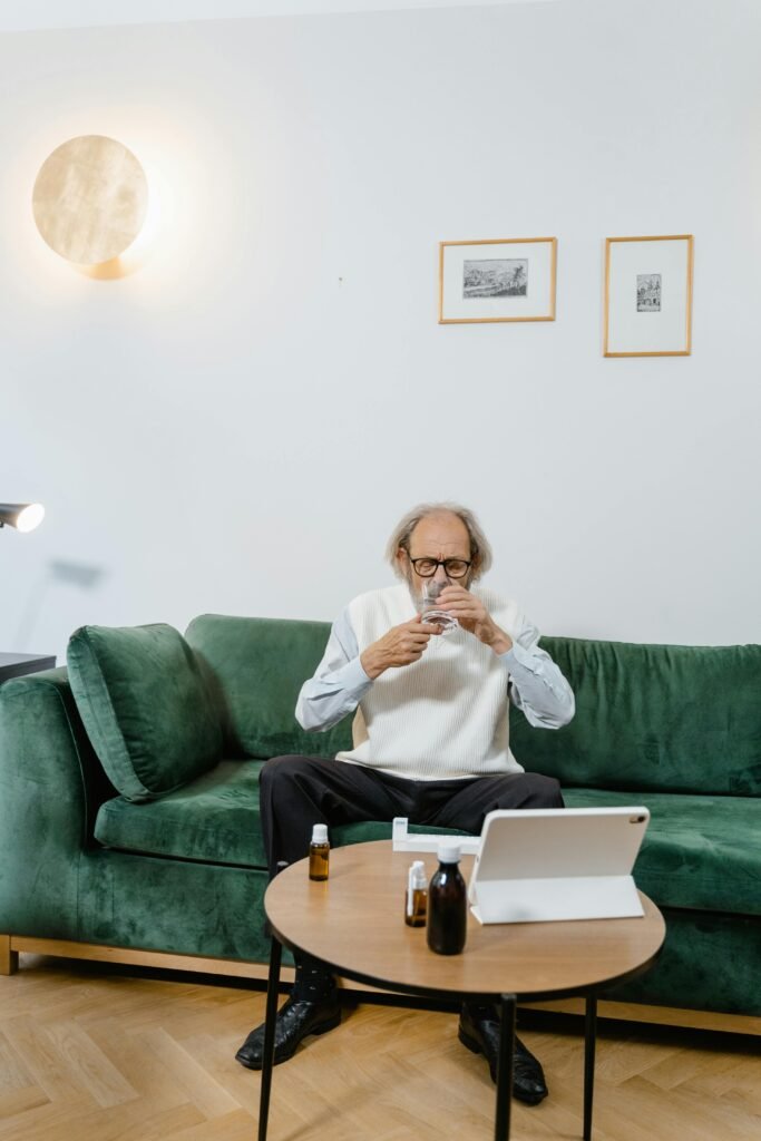 Elderly man in living room setting taking medication during virtual healthcare consultation.